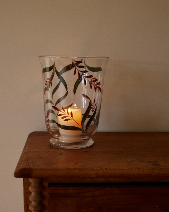 The Botanical Candle Holder, featuring decorative leaves on a glass hurricane vase, adds charm as it illuminates a wooden surface against a plain beige wall.
