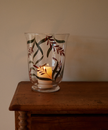 The Botanical Candle Holder, featuring decorative leaves on a glass hurricane vase, adds charm as it illuminates a wooden surface against a plain beige wall.