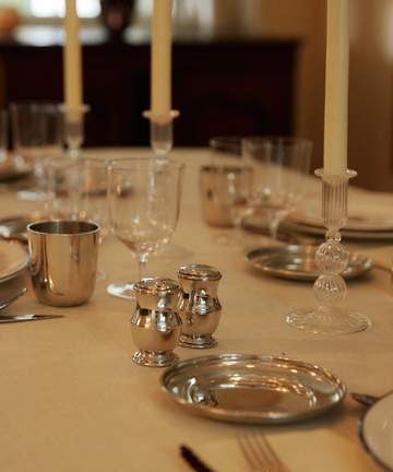 A formal table with silver plates, glasses, metal cups, the Evie Salt & Pepper Shaker, tall candles, and elegant tableware on a cream tablecloth.