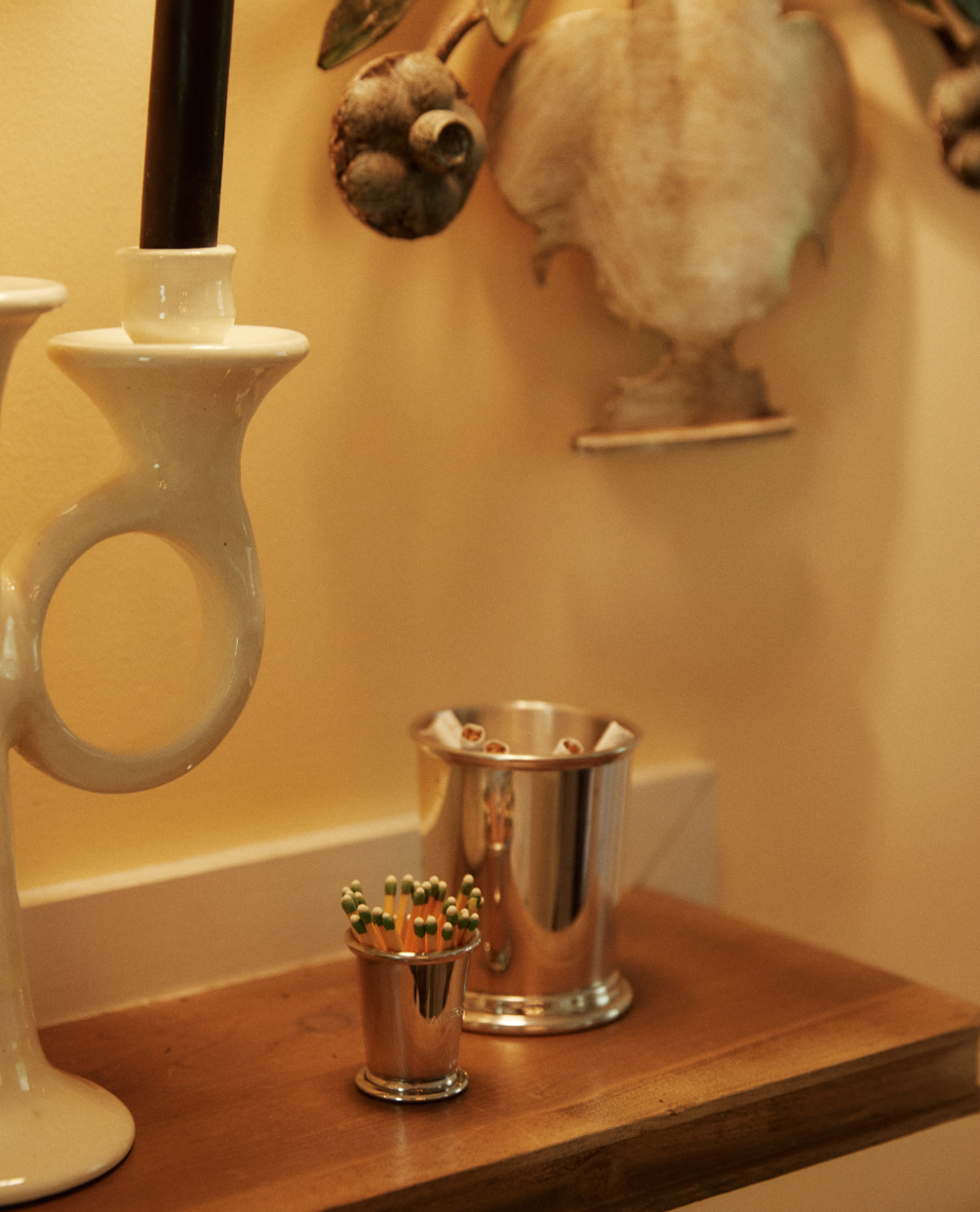 A wooden shelf holds a white ceramic candlestick, the Jude Match Cup & Striker with green-tipped matches, and a silver cup of pencils against a yellow decorative wall.