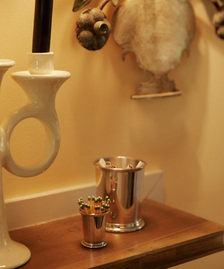A wooden shelf holds a white ceramic candlestick, the Jude Match Cup & Striker with green-tipped matches, and a silver cup of pencils against a yellow decorative wall.