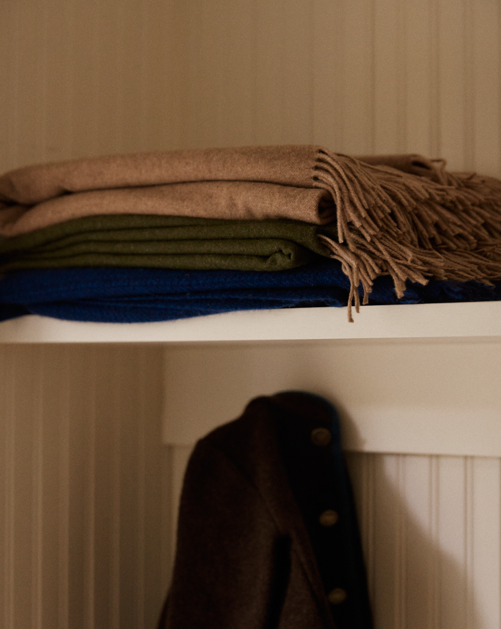 Three Ulisse Throw blankets—a neutral beige with fringe, a green merino wool, and a blue—are stacked on a white shelf above a dark coat against a paneled wall.