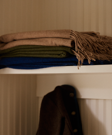 Three Ulisse Throw blankets—a neutral beige with fringe, a green merino wool, and a blue—are stacked on a white shelf above a dark coat against a paneled wall.