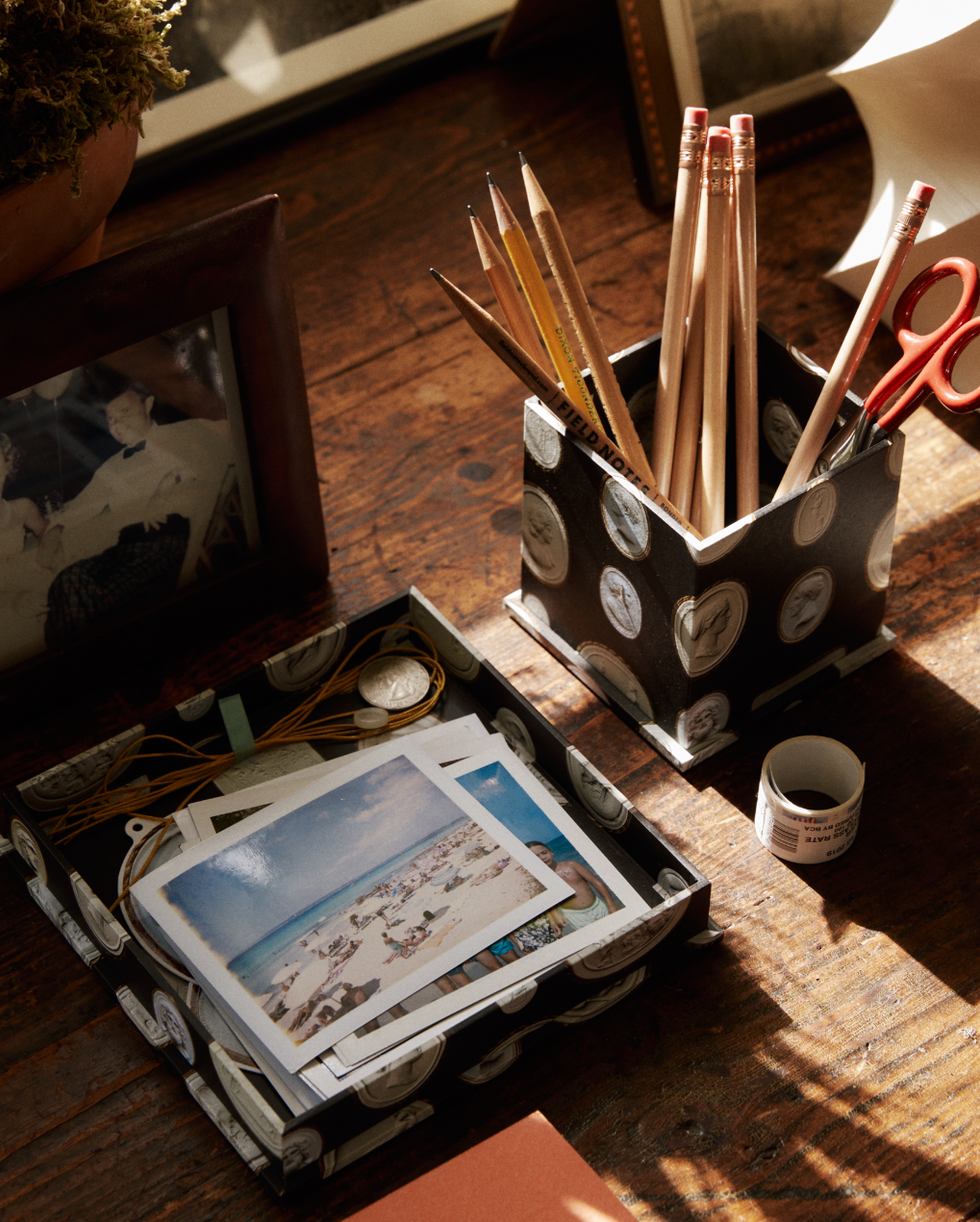 A desktop featuring a photo, a container of pencils and scissors, a small roll of tape, and a watch and postcards neatly organized in the Intaglio Nesting Tray on top of an archival book board covering the wooden surface.