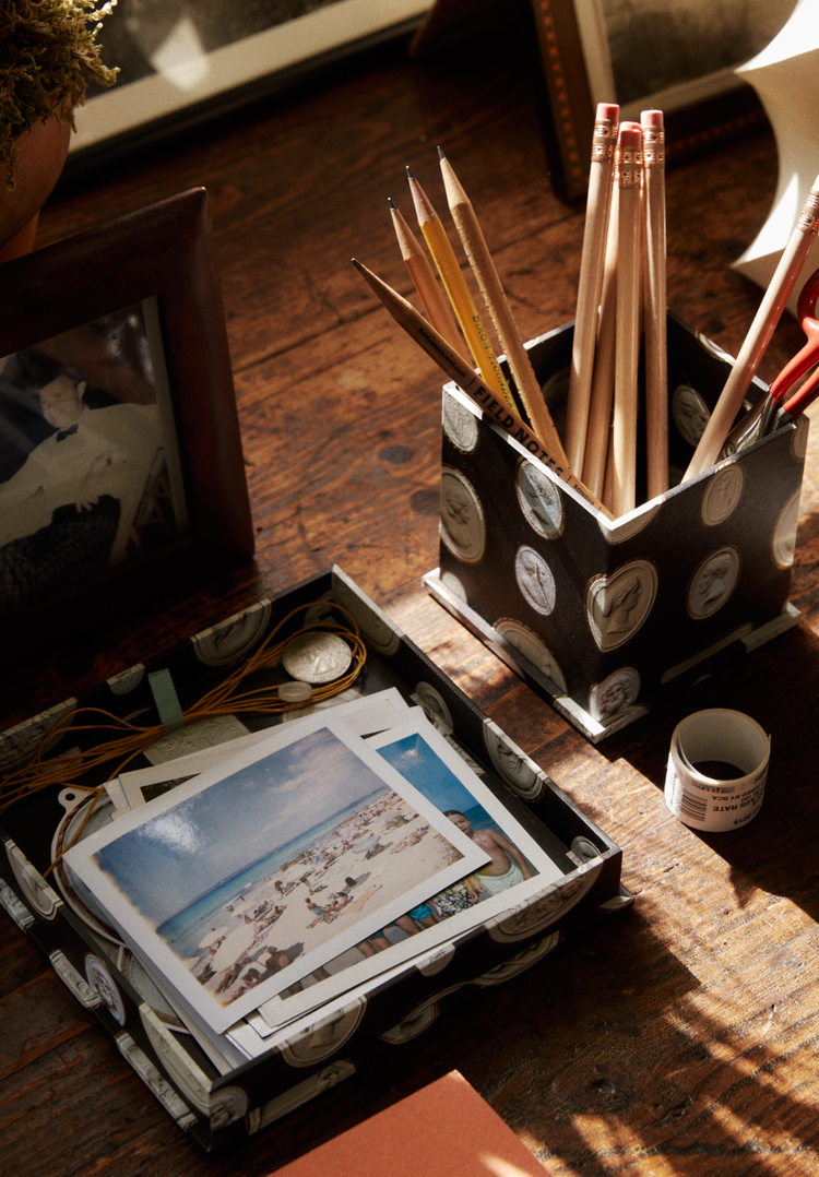 A desktop featuring a photo, a container of pencils and scissors, a small roll of tape, and a watch and postcards neatly organized in the Intaglio Nesting Tray on top of an archival book board covering the wooden surface.