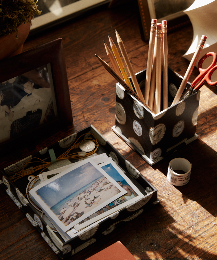 A desktop featuring a photo, a container of pencils and scissors, a small roll of tape, and a watch and postcards neatly organized in the Intaglio Nesting Tray on top of an archival book board covering the wooden surface.
