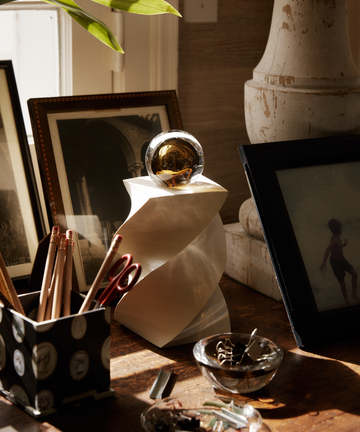 A Pallino Paperweight, featuring a unique glass sphere with a swirling amber interior, sits atop a white, twisted geometric pedestal on a desk.