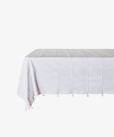 The Cordon Tablecloth, a white rectangular cotton cover with subtle vertical stripes and fringe tassels, is draped over a table against a plain white backgroundâ€”ideal for summer gatherings.