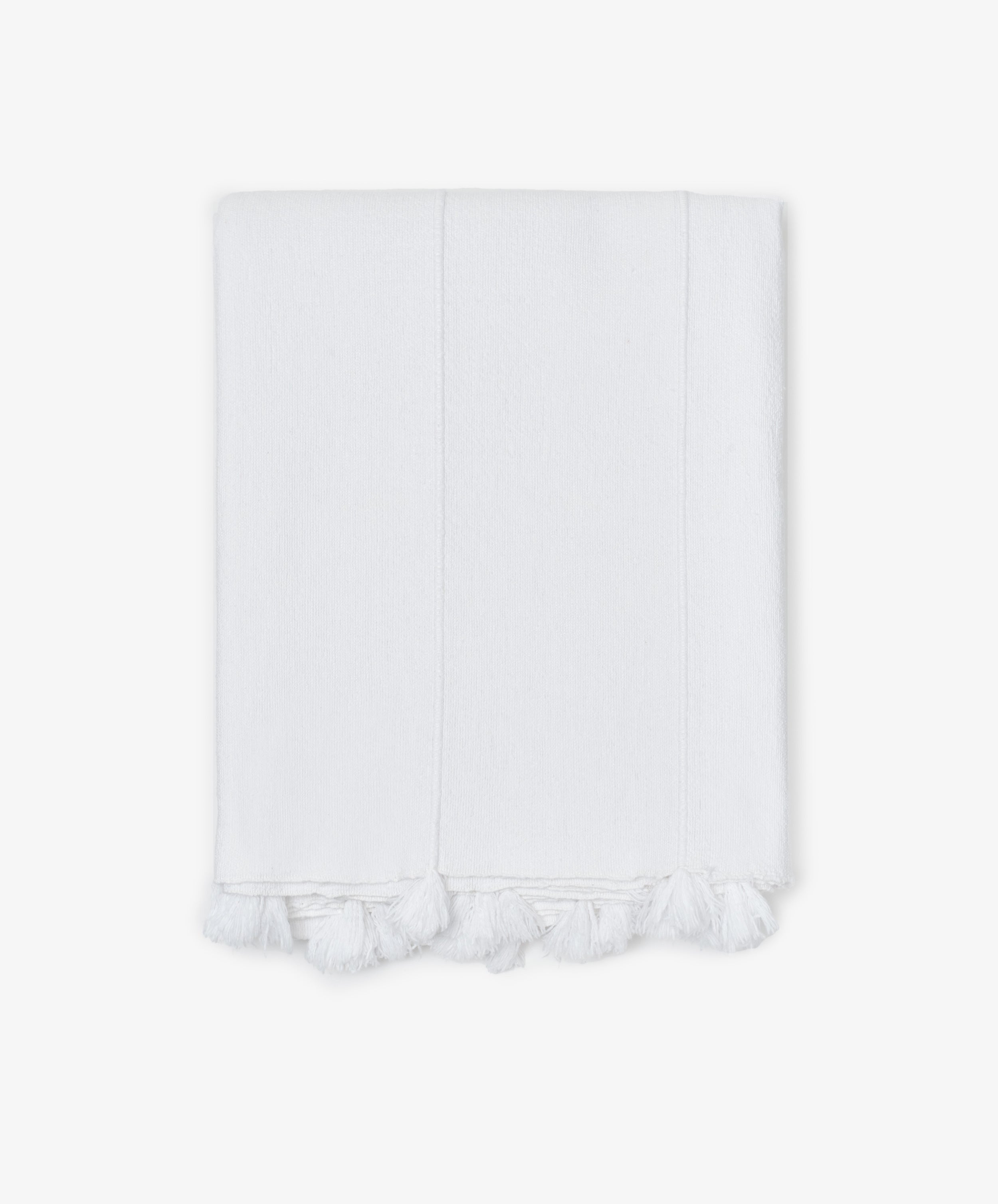 The Cordon Tablecloth is made from soft white cotton, featuring stitched lines and delicate fringe tassels along the edge, shown folded against a plain white background.