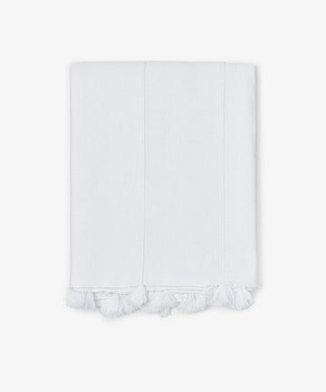 The Cordon Tablecloth is made from soft white cotton, featuring stitched lines and delicate fringe tassels along the edge, shown folded against a plain white background.