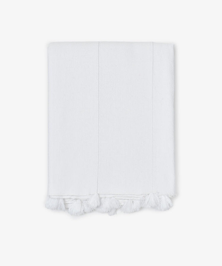 The Cordon Tablecloth is made from soft white cotton, featuring stitched lines and delicate fringe tassels along the edge, shown folded against a plain white background.
