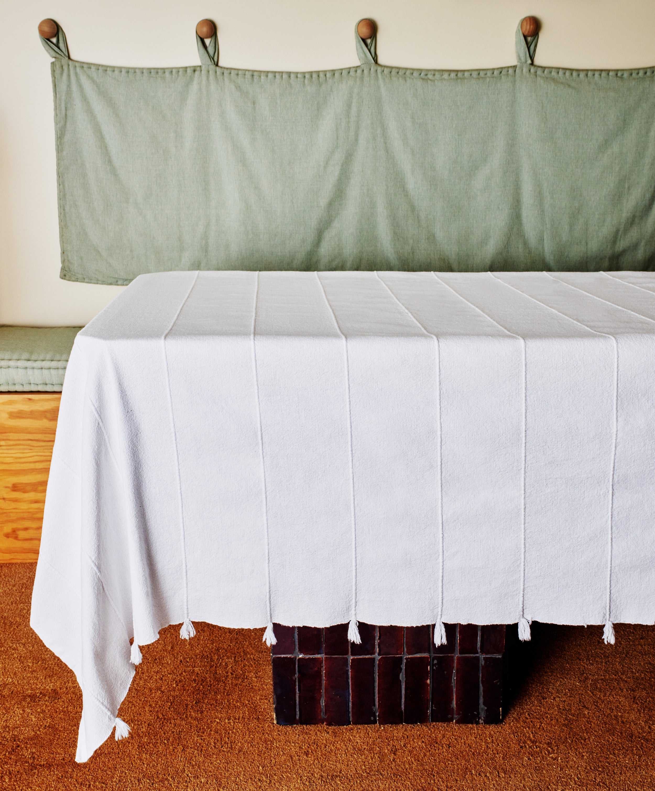 A table draped with the Cordon Tablecloth sits before a cushioned bench with a green fabric backrest, wooden pegs above, and rests on a brown carpet—ideal for relaxed summer gatherings.