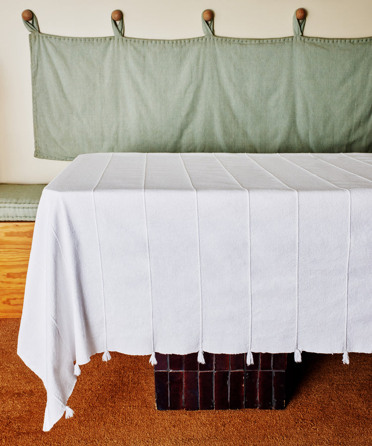 A table draped with the Cordon Tablecloth sits before a cushioned bench with a green fabric backrest, wooden pegs above, and rests on a brown carpet—ideal for relaxed summer gatherings.