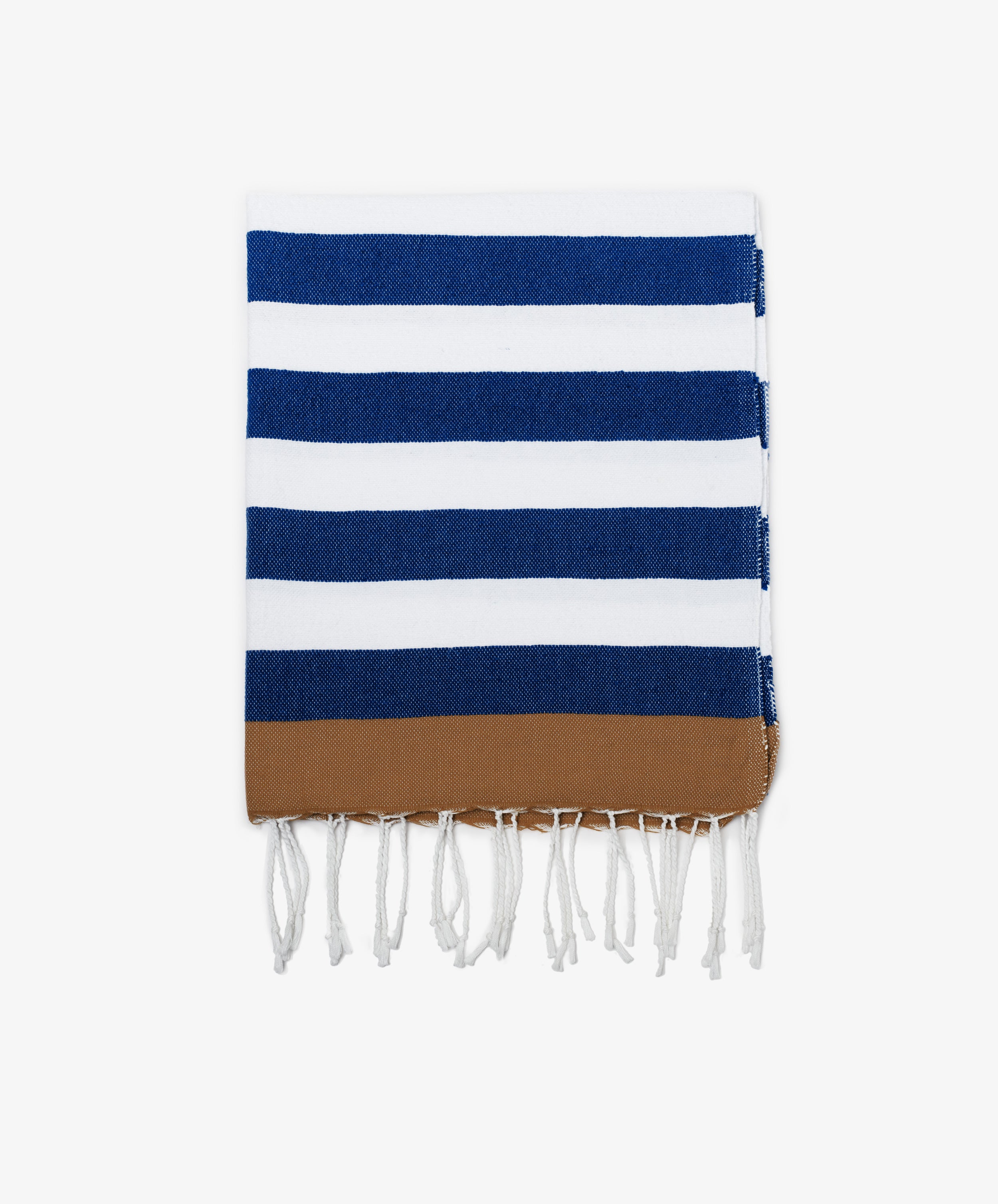 The La Plage Towel is a folded, lightweight towel featuring blue and white horizontal stripes, a brown band at one end, and white fringed tassels along the edge.