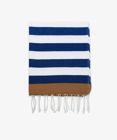 The La Plage Towel is a folded, lightweight towel featuring blue and white horizontal stripes, a brown band at one end, and white fringed tassels along the edge.