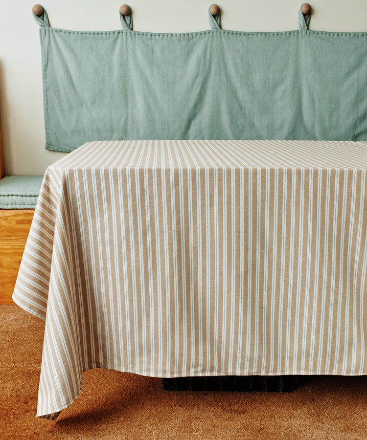 The Martin Striped Tablecloth, in beige and white cotton, covers a rectangular table set by a green fabric wall with coat hooks, showcasing the classic charm of durable table linens.
