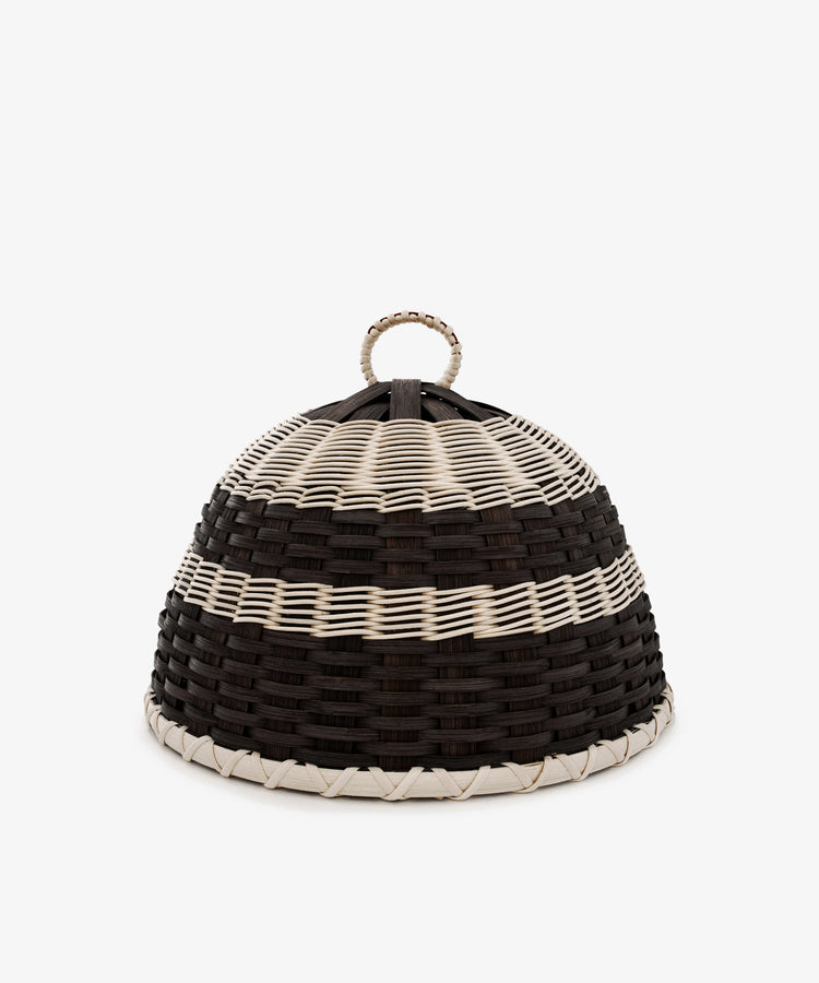 The Folk Fleur Cloche is a dome-shaped, handwoven basket featuring black, white, and brown horizontal stripes and a loop handle on top. Perfect as a stylish cheese cover for outdoor entertaining, it is set against a plain background.