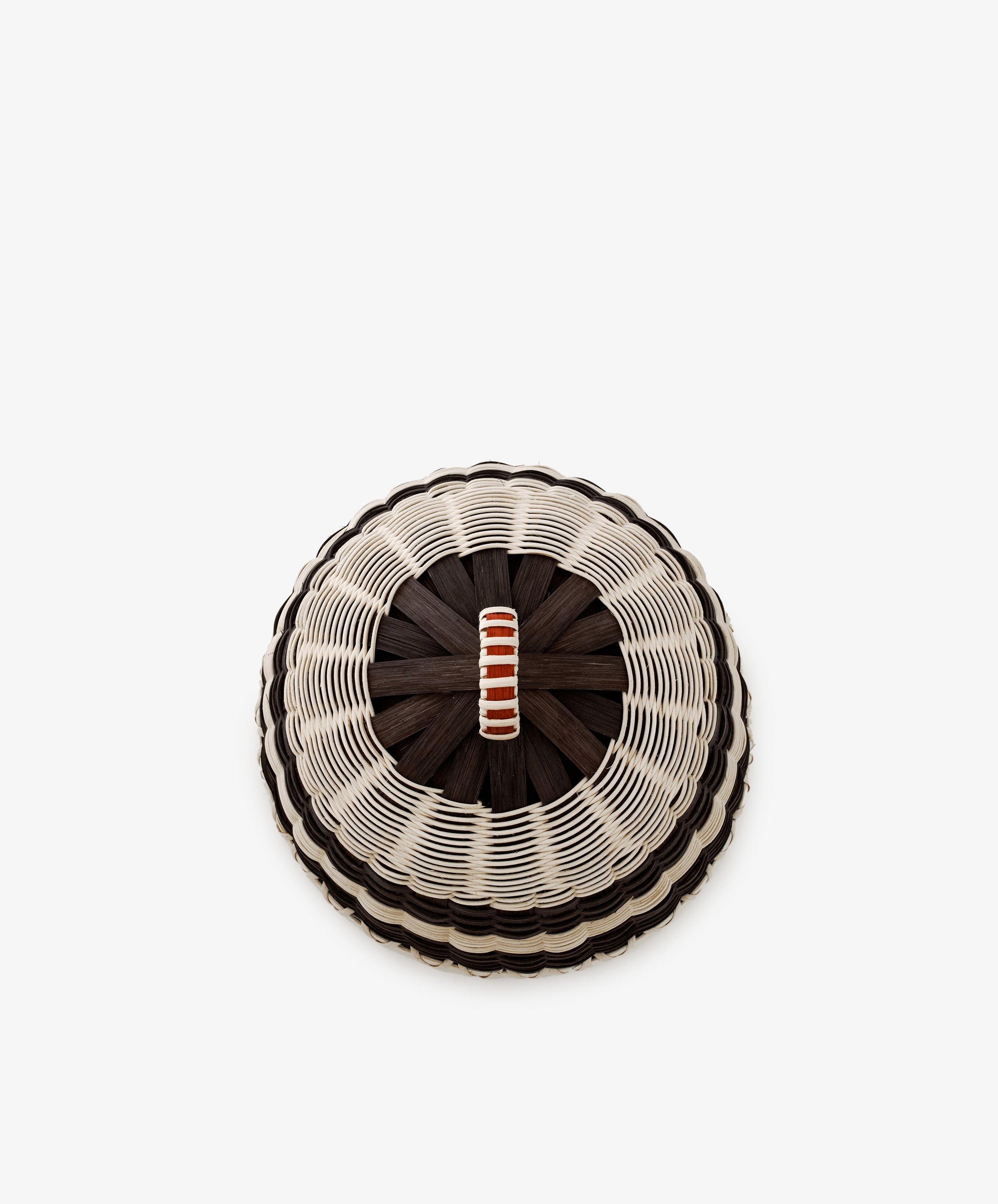 Showcased against a white background, the Folk Fleur Cloche features a round woven lid with a striking black center, white and dark brown radial weaving, and a small orange-and-white striped handle. Handwoven and ideal as a cheese cover, it’s perfect for outdoor entertaining.