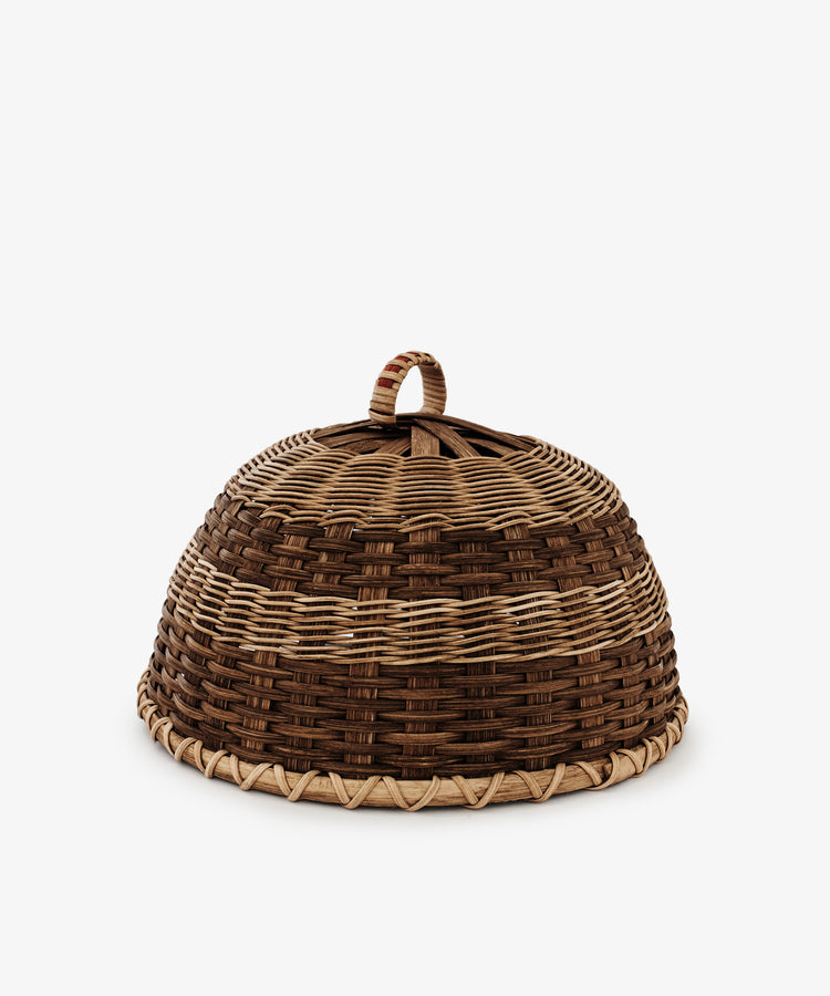 The Folk Fleur Cloche is a dome-shaped, handwoven piece with a top handle and alternating light and dark brown bands—perfect for outdoor entertaining.