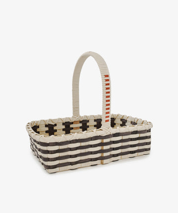 The Folk Fleur Hand Basket is a rectangular woven basket with a tall handle, showcasing alternating black, white, and beige bands and a small orange accent on the handle. Set against a white background, this stylish market basket is perfect for carrying essentials or adding an elegant touch to your storage solutions.
