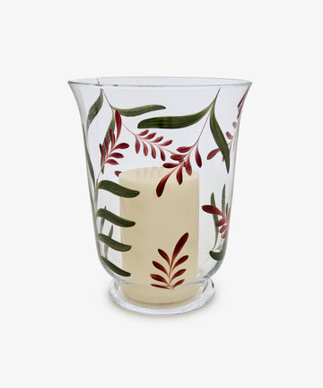 The Botanical Candle Holder is a clear glass hurricane featuring a decorative leaf design with painted green leaves and red branches, and includes an unlit white pillar candle—making it perfect as a striking table centerpiece.