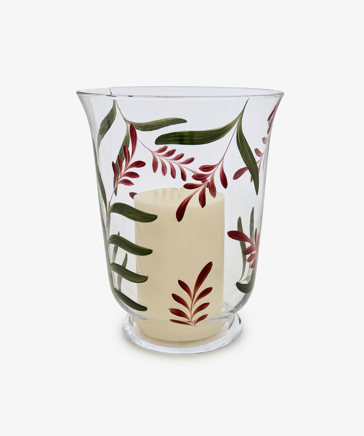 The Botanical Candle Holder is a clear glass hurricane featuring a decorative leaf design with painted green leaves and red branches, and includes an unlit white pillar candle—making it perfect as a striking table centerpiece.