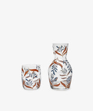 The Botanical Bedside Carafe and Glass Set features orange and blue leaf patterns hand-painted on clear glass, elegantly displayed on a plain white background.