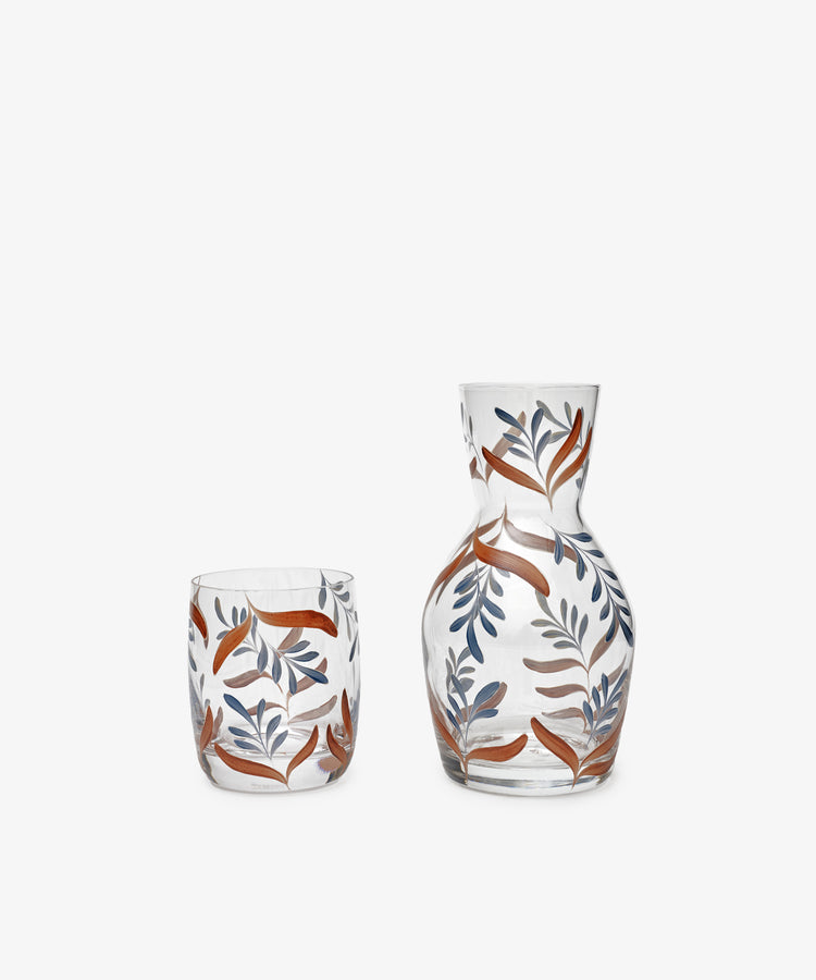 The Botanical Bedside Carafe and Glass Set features orange and blue leaf patterns hand-painted on clear glass, elegantly displayed on a plain white background.