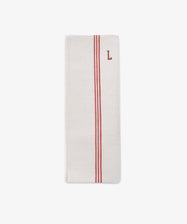The French Vintage Tea Towel is a folded white cloth napkin with two parallel red stripes and a red embroidered letter L near the top right corner, evoking classic vintage linen tea towels.