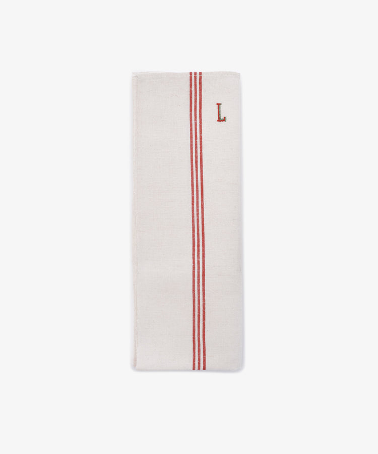 The French Vintage Tea Towel is a folded white cloth napkin with two parallel red stripes and a red embroidered letter L near the top right corner, evoking classic vintage linen tea towels.