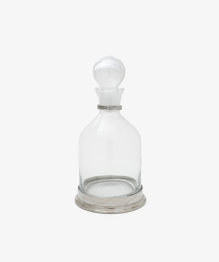 The Alec Decanter features clear glass with a pewter collar and round stopper, set on a silver-hued metal base, displayed against a plain white background.