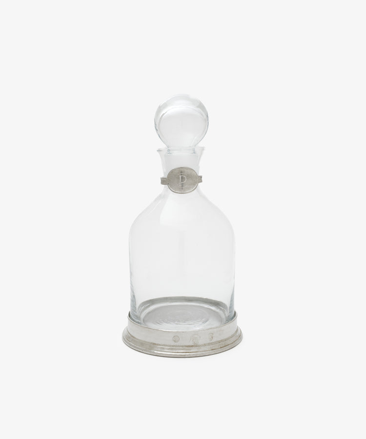 The Alec Decanter is a clear glass decanter with a round pewter stopper, pewter collar and base, all engraved with decorative markings and the letter P.