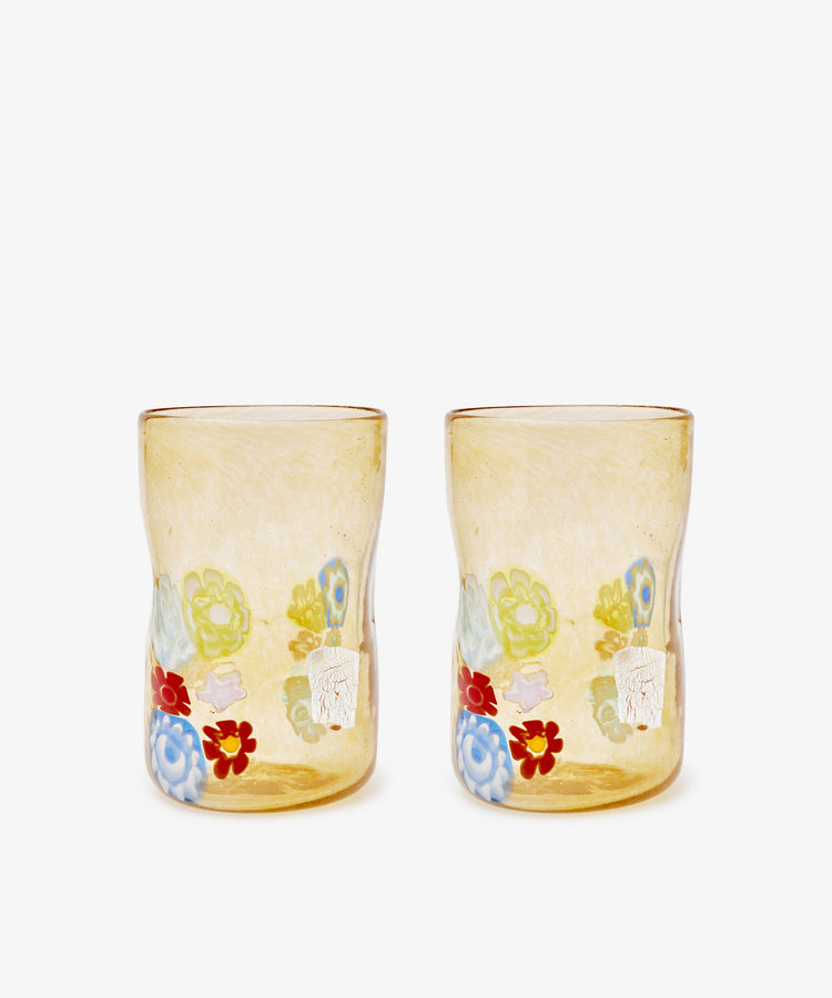 The Muranesi Glass Set of 2 features yellow handblown tumblers with a unique shape, each adorned with vibrant red, blue, green, and white floral patterns near the base—classic Murano-inspired elegance.