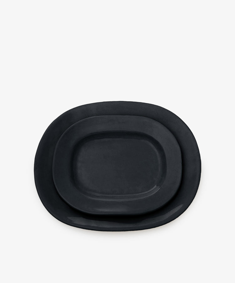 A large black, matte-finish, rectangular platter is stacked with a smaller plate on a plain white background.