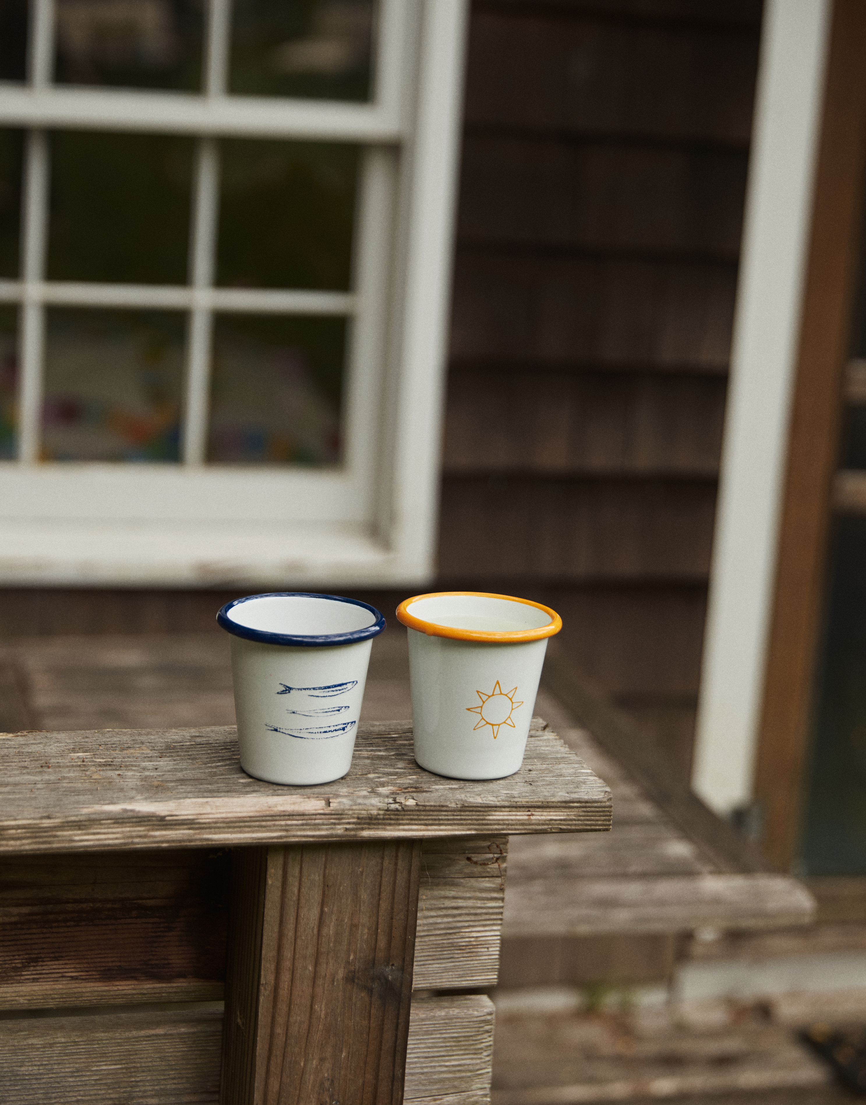 The Il Sole Enamel Cup, Set of 2, sits on a weathered ledge—one cup with blue lines and rim, the other with a sun motif and orange rim. A house window and siding appear in the background, evoking summer vibes.