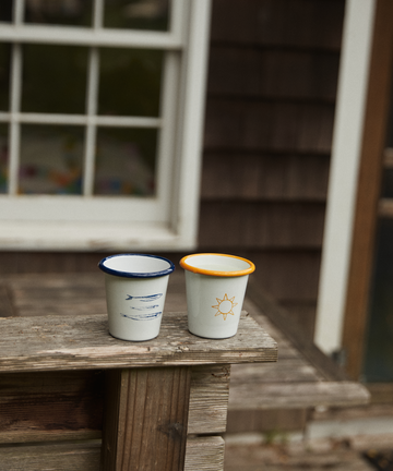The Il Sole Enamel Cup, Set of 2, sits on a weathered ledge—one cup with blue lines and rim, the other with a sun motif and orange rim. A house window and siding appear in the background, evoking summer vibes.