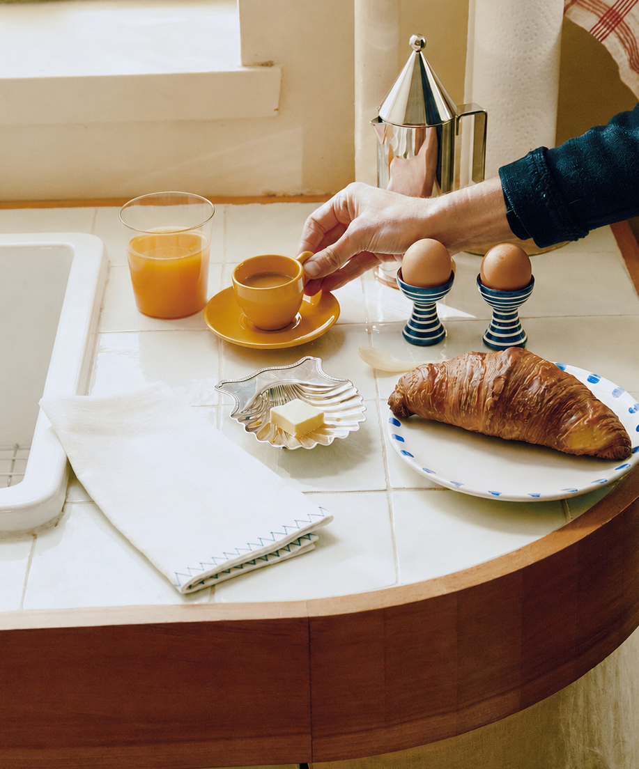 Person preparing breakfast at a kitchen counter with coffee, croissant, and eggs.