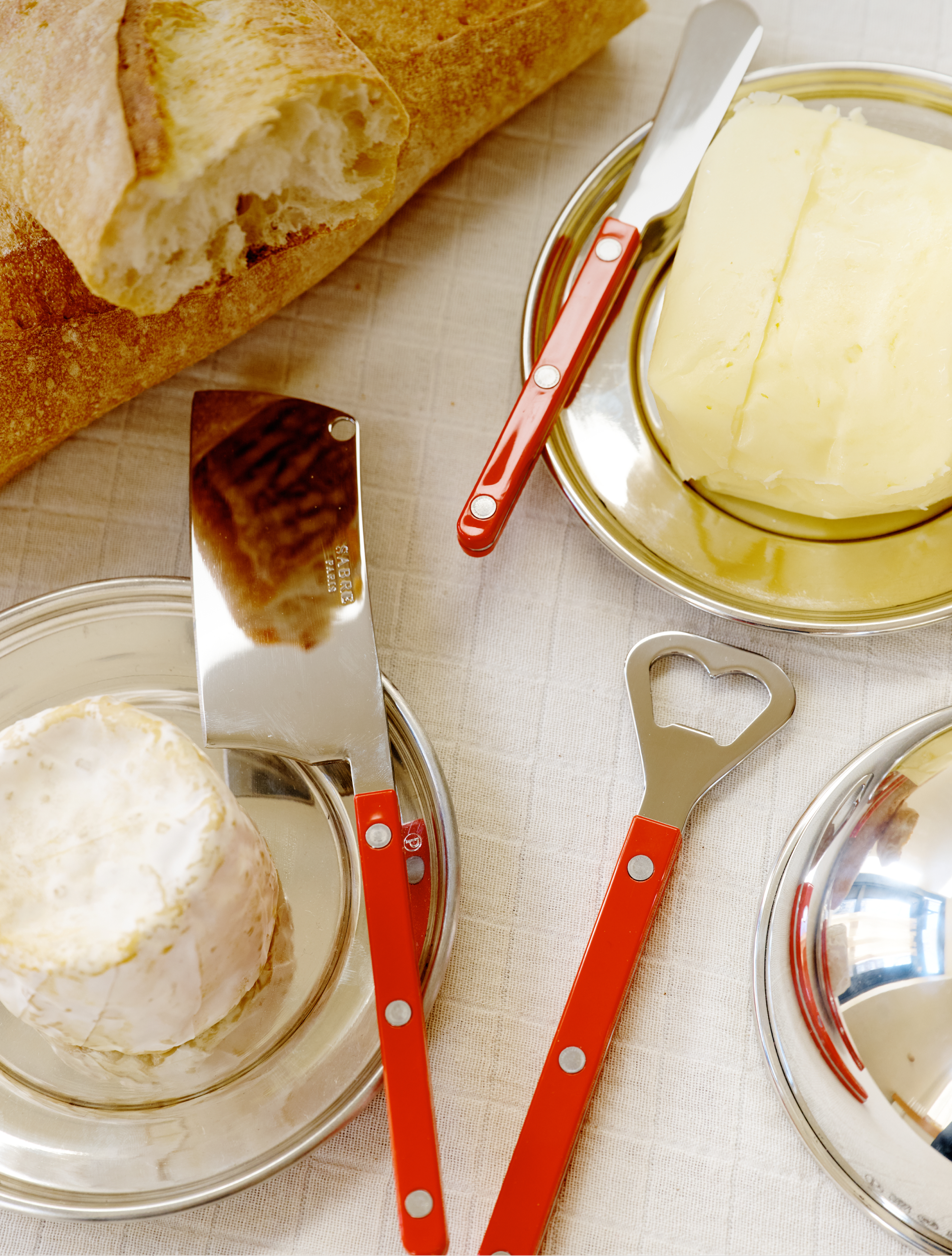 The Good Bits Bundle—featuring a loaf of bread, round cheese, and slab butter—is arranged with a red-handled cheese cleaver and butter knife on plates atop a white tablecloth.