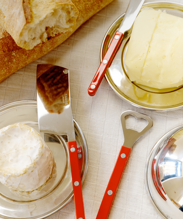 The Good Bits Bundle—featuring a loaf of bread, round cheese, and slab butter—is arranged with a red-handled cheese cleaver and butter knife on plates atop a white tablecloth.