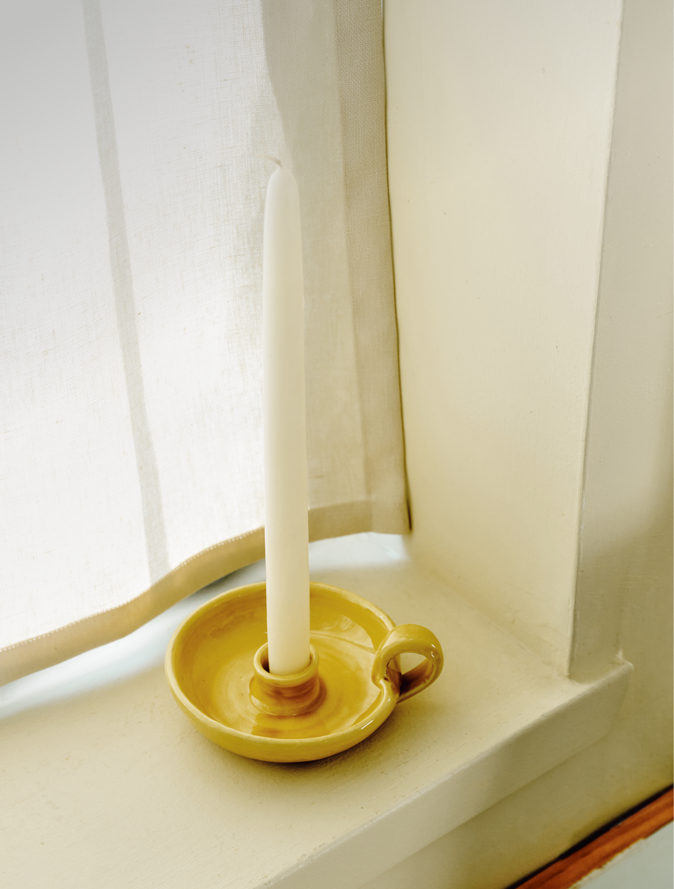 A white tapered candle from the Sparks Flying Bundle stands upright in a yellow ceramic candleholder on a windowsill next to a white curtain.
