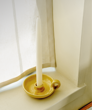 A white tapered candle from the Sparks Flying Bundle stands upright in a yellow ceramic candleholder on a windowsill next to a white curtain.