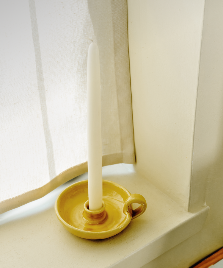 A white tapered candle from the Sparks Flying Bundle stands upright in a yellow ceramic candleholder on a windowsill next to a white curtain.