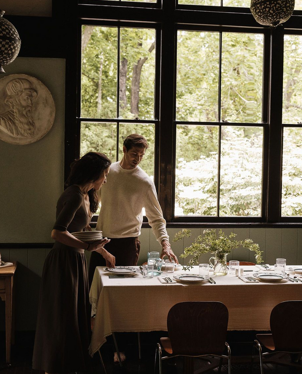 A man and a woman standing beside a dining table in a rustic room with large black-framed windows looking out onto a green wooded scene. The man wears a cream knit sweater and dark trousers, and the woman wears a dark brown dress while holding plates.
