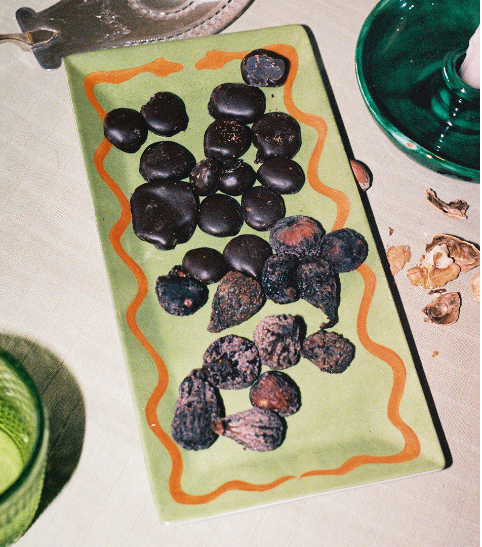 A rectangular Hand Painted Double Serpent Tray in green displays two types of snacks—round dark chocolate pieces and dried figs or nuts—and sits on a table alongside a green glass and walnut shells. The elegant tray showcases artisanal craftsmanship inspired by porcelain painters and third fire cooking techniques.