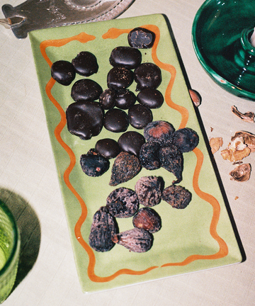 A rectangular Hand Painted Double Serpent Tray in green displays two types of snacks—round dark chocolate pieces and dried figs or nuts—and sits on a table alongside a green glass and walnut shells. The elegant tray showcases artisanal craftsmanship inspired by porcelain painters and third fire cooking techniques.