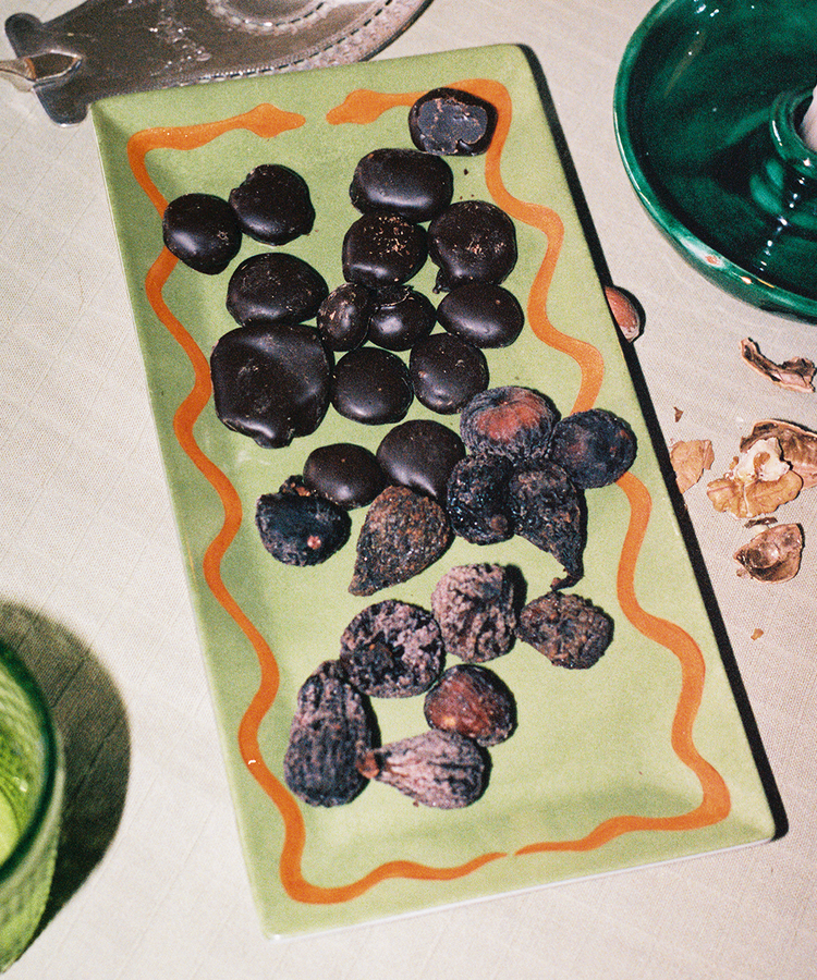 A rectangular Hand Painted Double Serpent Tray in green displays two types of snacks—round dark chocolate pieces and dried figs or nuts—and sits on a table alongside a green glass and walnut shells. The elegant tray showcases artisanal craftsmanship inspired by porcelain painters and third fire cooking techniques.