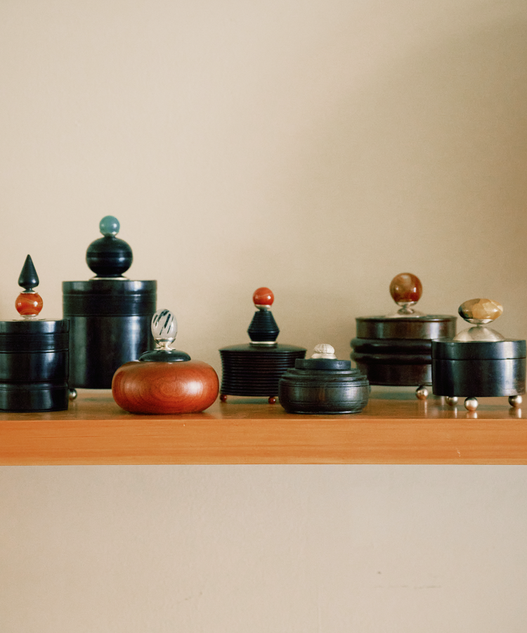 The Curiosity Box, a decorative wooden container with a unique knob lid, sits arranged on a light wooden shelf against a plain beige wall.