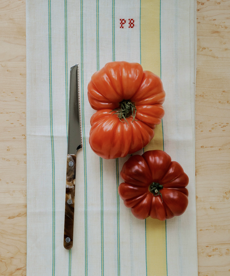 Two large heirloom tomatoes and a Tomato Knife sit on a striped kitchen towel with red 
