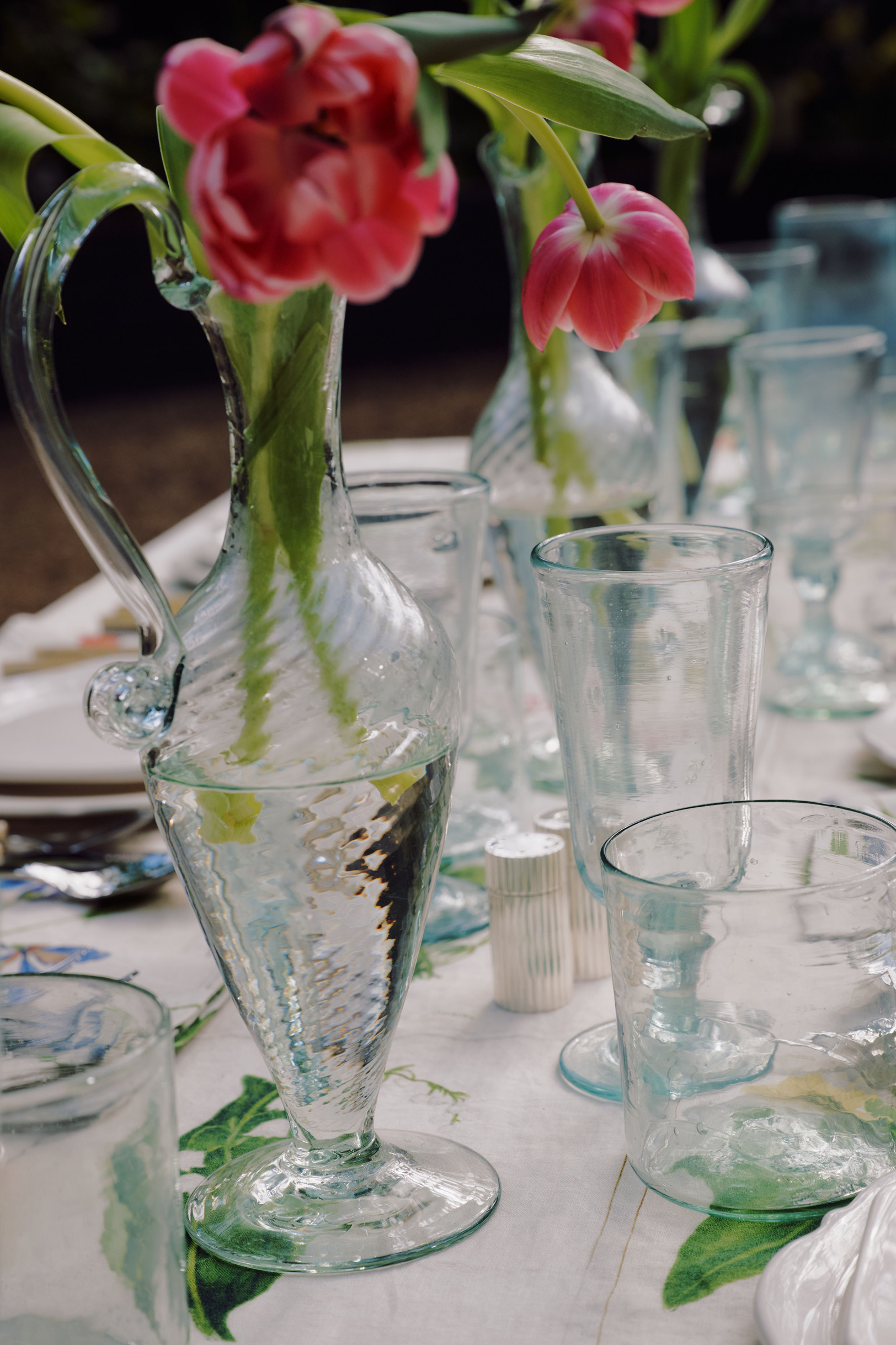 The Alba Vase, a clear glass pitcher holding pink tulips, stands surrounded by empty hand-blown vases on a decorated outdoor dining table.