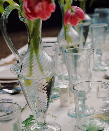 The Alba Vase, a clear glass pitcher holding pink tulips, stands surrounded by empty hand-blown vases on a decorated outdoor dining table.